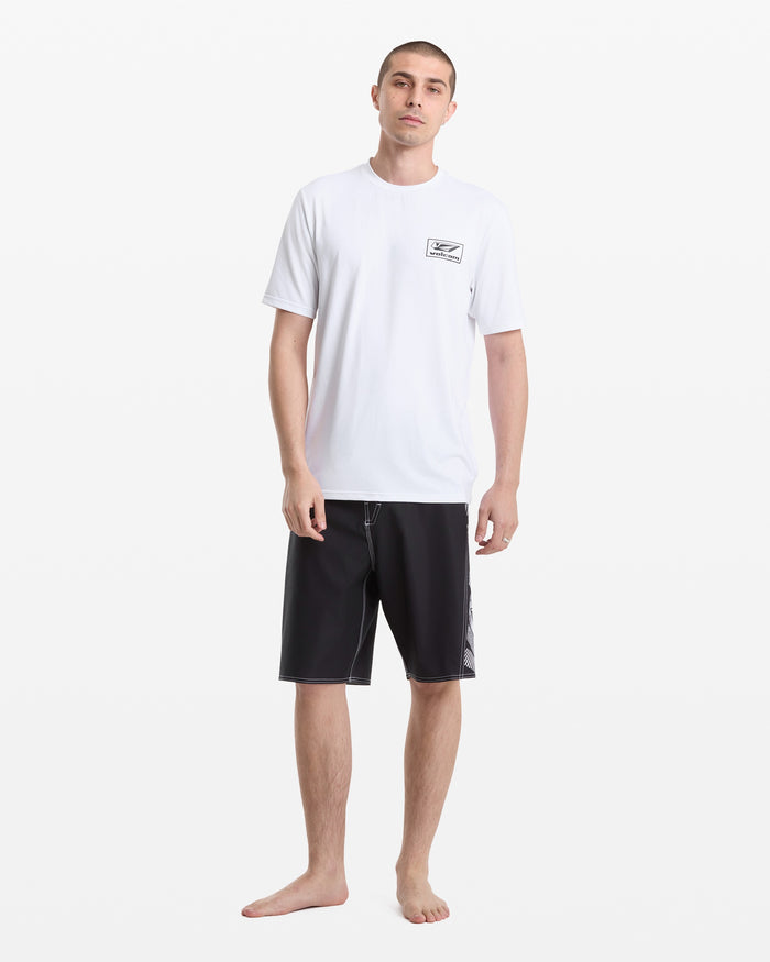 A man stands barefoot against a white background, wearing the Volcom Men's Modstone Loose Short Sleeve Surf Tee with a small chest logo and black knee-length shorts. The tee offers a relaxed fit and loose short sleeves for comfort.