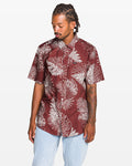 A man with braided hair stands against a white background, wearing the Volcom Men's Monsterious Short Sleeve Shirt—a burgundy button-up featuring an all-over white tropical leaf print. He looks down to his left with one hand by his side.