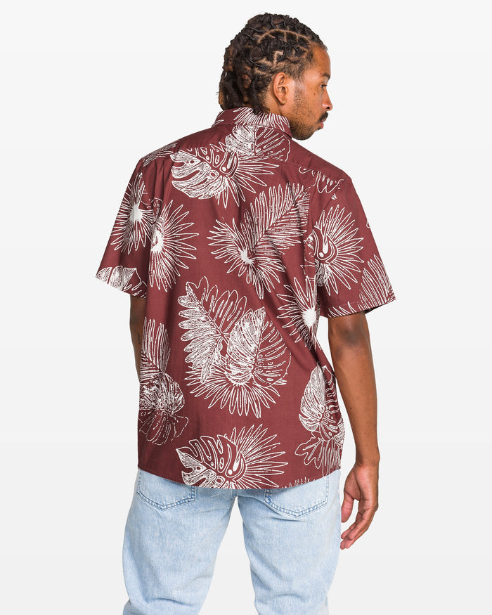 A man with braided hair wears the Volcom Men's Monsterious Short Sleeve Shirt—a cotton viscose blend with white tropical leaf print on maroon—and light blue jeans. He stands with his back to the camera, looking over his shoulder.