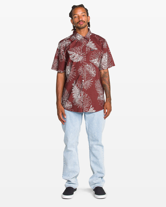 A man stands facing forward, wearing the Volcom Men's Monsterious Short Sleeve Shirt—a maroon cotton viscose blend with a white tropical leaf print—paired with light blue jeans and black slip-on shoes against a plain white background.