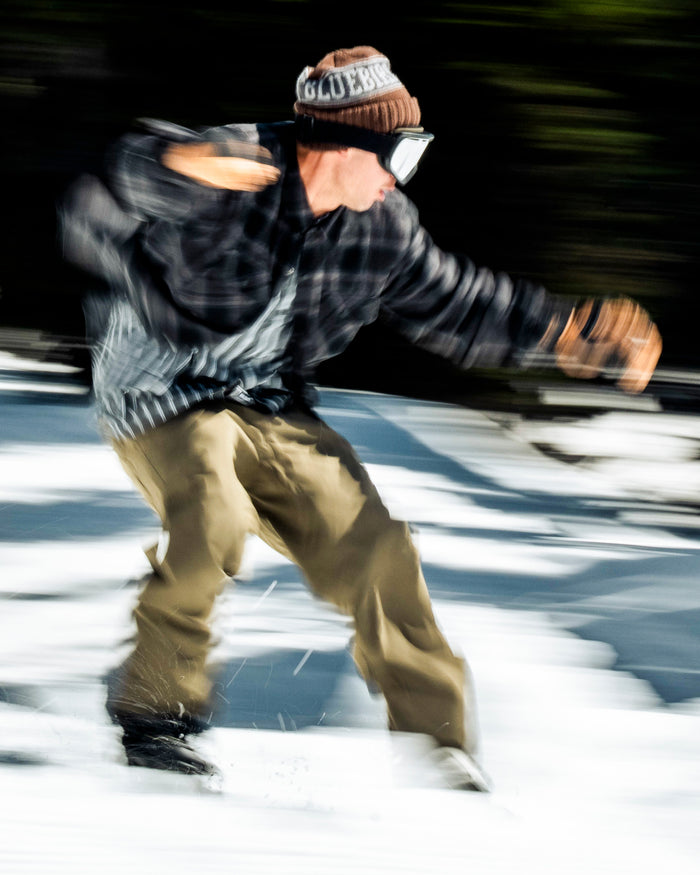 A snowboarder speeding across the snow, dressed in Volcom Men’s Nailer Pants in tan, paired with a plaid jacket, goggles, and a brown beanie; motion blur highlights the sense of rapid movement.
