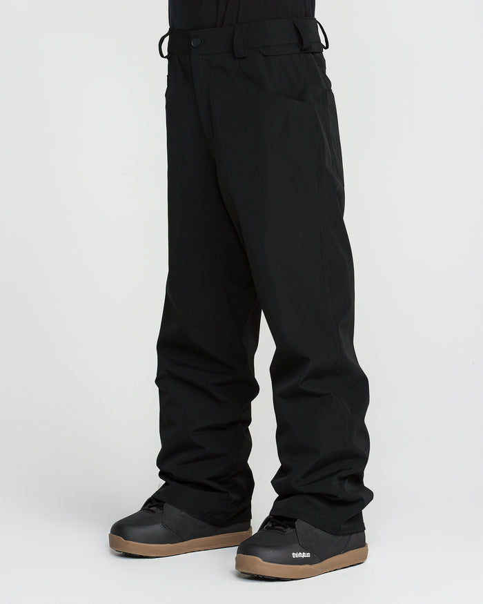 A person wearing black Volcom Men’s Nailer Pants, crafted from recycled materials, and black snow boots with brown soles, stands against a plain white background.