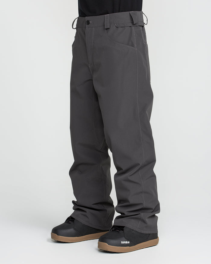 A person wearing Volcom Men’s Nailer Pants in dark gray and black snow boots with brown soles, standing against a plain white background. Only the lower half of the body is visible.