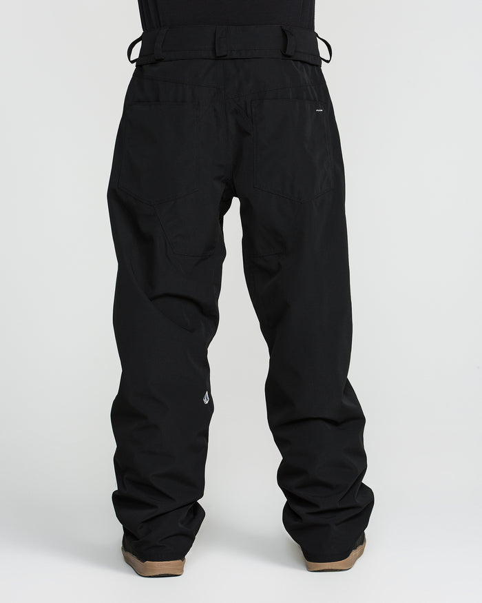A person is shown from the back against a plain white background wearing Volcom Men’s Nailer Pants—loose-fitting black waterproof pants made from recycled materials—and brown shoes.