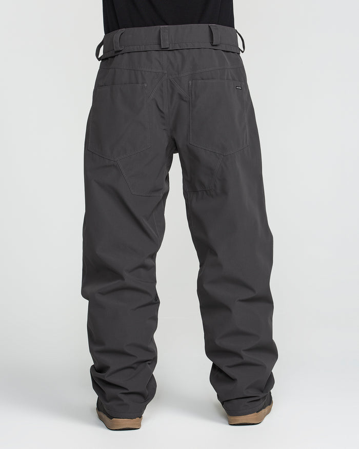 A person is shown from the back against a plain white background wearing Volcom Men’s Nailer Pants in dark gray, which are loose-fitting, waterproof, and crafted from recycled fabric. The pants feature large back pockets, sturdy belt loops, and are paired with tan shoes.