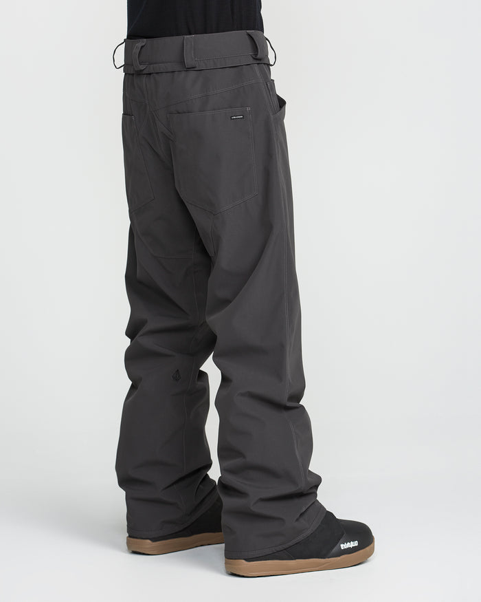 A person stands facing away against a plain white background, wearing Volcom Men’s Nailer Pants—loose-fitting and made from dark gray recycled fabric—paired with black shoes featuring brown soles.