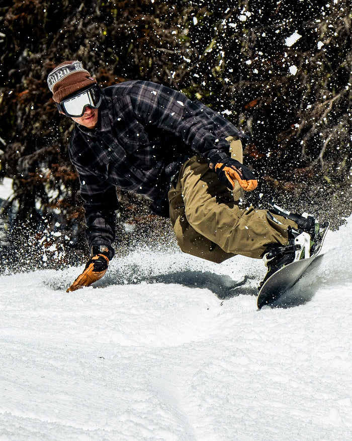 Wearing a brown beanie, goggles, plaid jacket, and Volcom Men’s Nailer Pants in khaki, a snowboarder carves quickly through fresh snow—kicking up a spray with trees in the background.