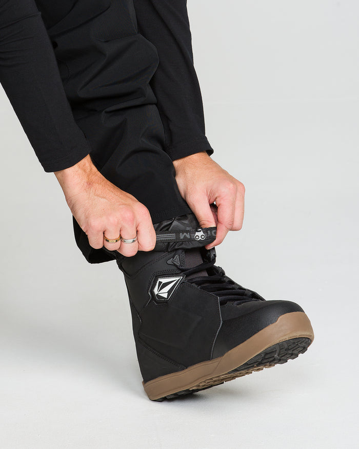 A person wearing Volcom Men’s Nailer Pants, crafted from recycled materials, adjusts the strap on a black snowboard boot with a brown sole. The image highlights their hands and the boot against a light background.