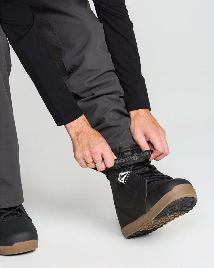 A person adjusts the strap on a black snow boot over Volcom Men’s Nailer Pants in gray, preparing for outdoor activity. The photo focuses on the hands, the boot, and part of the lower legs against a plain background.