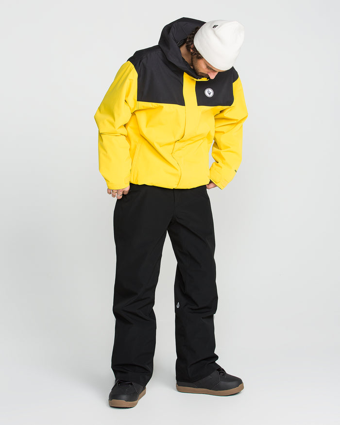 A person wearing a yellow and black jacket, black Volcom Men’s Nailer Pants made from recycled materials, black shoes, and a white beanie stands looking down against a plain white background.
