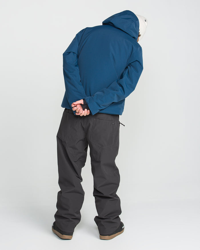 A person is seen from behind against a plain white background, wearing Volcom Men’s Nailer Pants in a dark recycled fabric, a blue hooded jacket, and a white helmet with their hands clasped behind their back.
