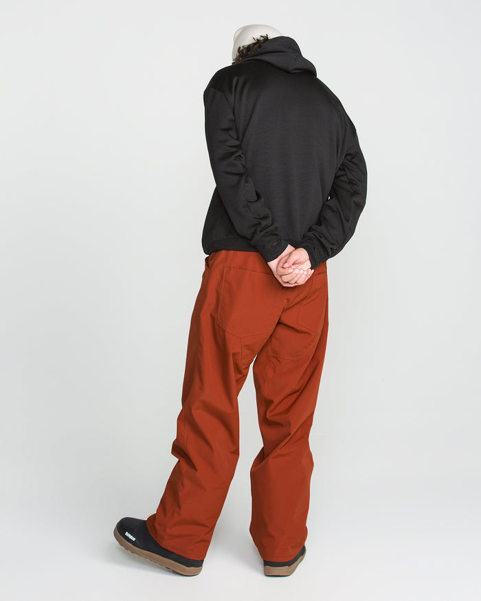 A person stands with their back to the camera against a plain white background, wearing a black hoodie, black shoes, and rust-colored Volcom Men’s Nailer Pants made from recycled fabric, with their hands clasped behind their back.