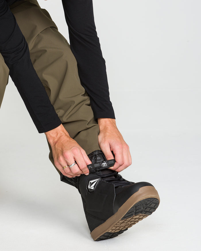 A person wearing Volcom Men’s Nailer Pants in olive green and a black long-sleeve shirt is tightening the laces on a black snowboarding boot with a brown sole.