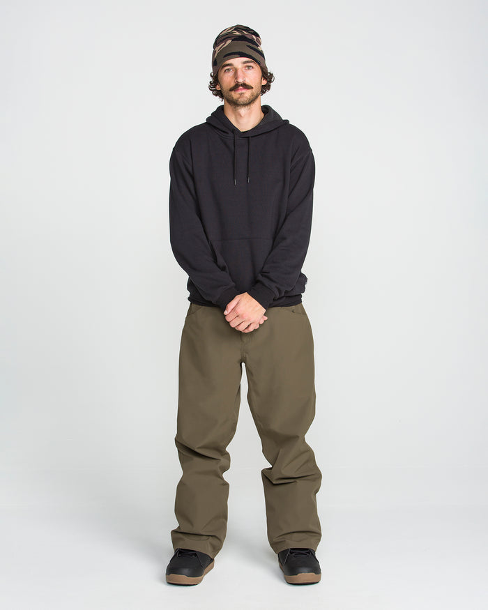 A person stands against a white background, wearing Volcom Men’s Nailer Pants in olive green with a baggy fit, paired with a black hoodie, black shoes, and a patterned beanie, hands clasped in front.