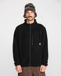 A man with a mustache wears the Volcom Men’s Nelson Sherpa Full Zip jacket, paired with brown pants and a camouflage beanie, standing against a plain white background.