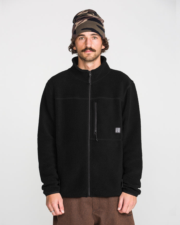 A man with a mustache wears the Volcom Men’s Nelson Sherpa Full Zip jacket, paired with brown pants and a camouflage beanie, standing against a plain white background.