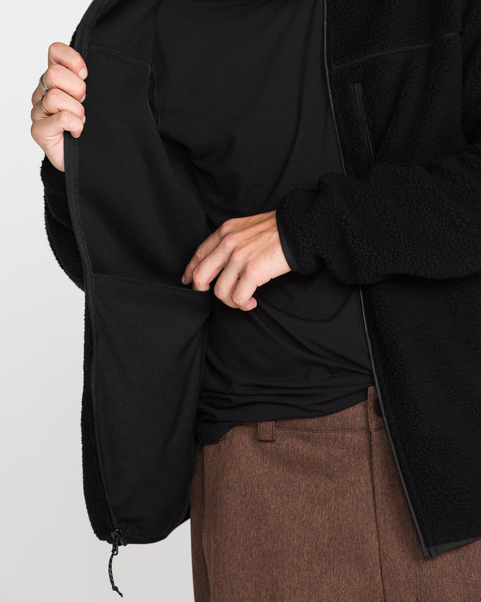 Wearing the Volcom Men’s Nelson Sherpa Full Zip in black, a person partially unzips the jacket to reveal an inner pocket, paired with a black shirt and brown pants. Only their torso and hands are visible.
