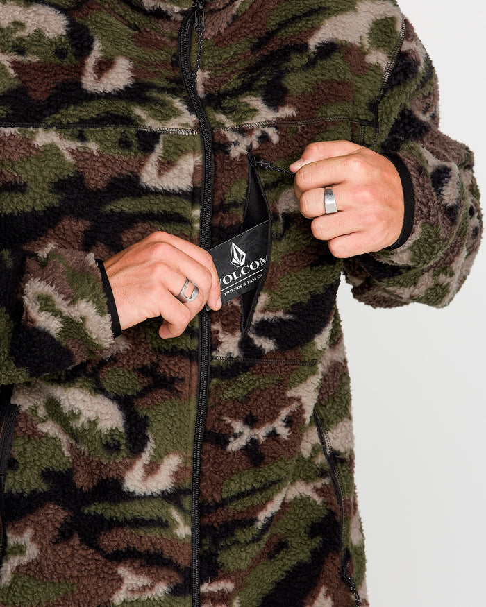 A person wearing the Volcom Men’s Nelson Sherpa Full Zip jacket in green and brown camo unzips a chest pocket to reveal the Volcom tag, with both hands visible and silver rings on their fingers.