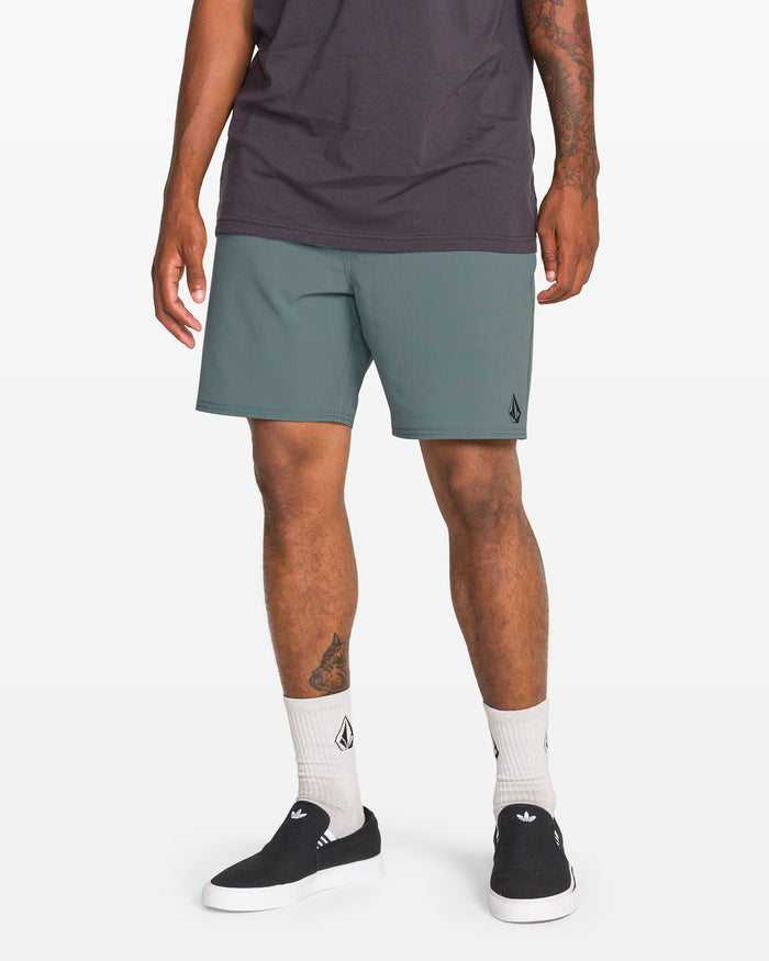 A person stands against a white background wearing a dark t-shirt, Volcom Men's Nomoly Elastic Waist Shorts 18" made from recycled polyester, white socks, and black slip-on sneakers. Tattoos are visible on their arm and leg.