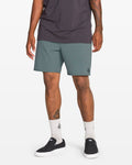 A person stands against a white background wearing a dark t-shirt, Volcom Men's Nomoly Elastic Waist Shorts 18" made from recycled polyester, white socks, and black slip-on sneakers. Tattoos are visible on their arm and leg.
