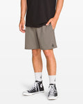 A person wearing a black t-shirt, Volcom Men's Nomoly Elastic Waist Shorts 18" in gray with a small logo, white socks, and black high-top sneakers stands with one hand in their pocket against a white background.