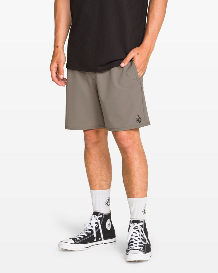 A person wearing a black t-shirt, Volcom Men's Nomoly Elastic Waist Shorts 18" in gray with a small logo, white socks, and black high-top sneakers stands with one hand in their pocket against a white background.
