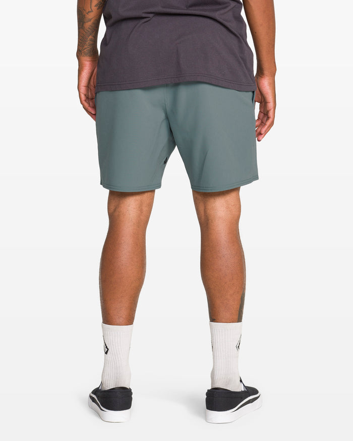 A person faces away from the camera, wearing a dark t-shirt and Volcom Men's Nomoly Elastic Waist Shorts 18" in teal-green, made from recycled polyester, with white crew socks and black slip-on sneakers against a white background.