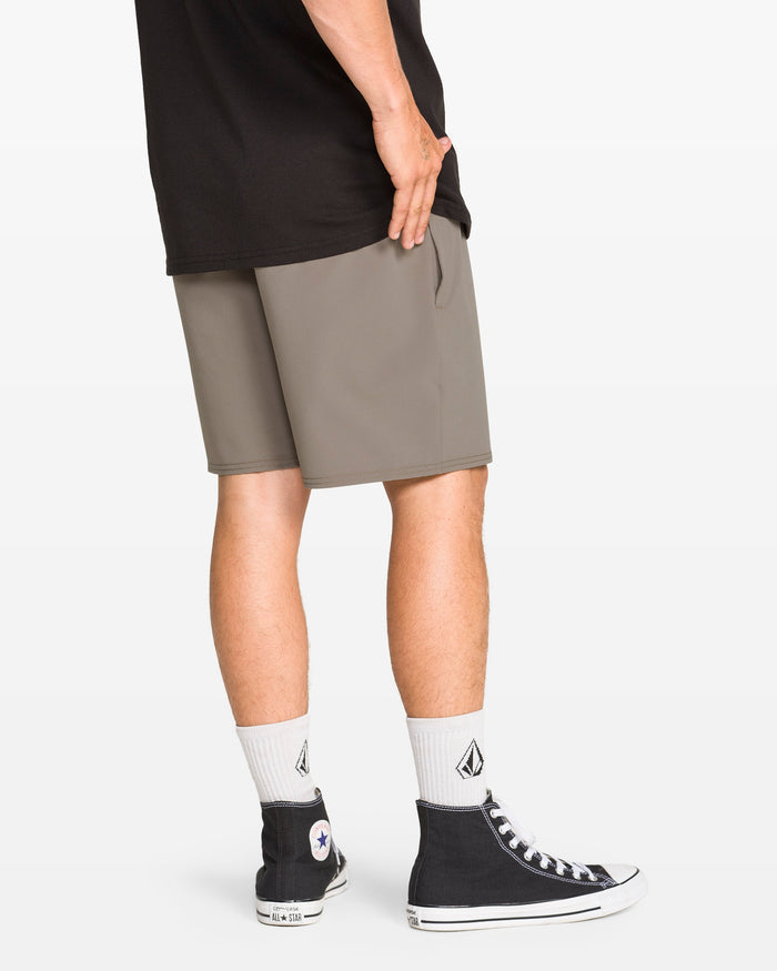 A person stands facing away on a white background, wearing a black t-shirt, Volcom Men's Nomoly Elastic Waist Shorts 18" in gray (4-way stretch, recycled polyester), white socks with a black logo, and black high-top Converse sneakers.