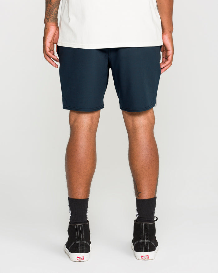 A back view of a person wearing a white shirt paired with Volcom Men's Nomoly Elastic Waist Shorts in dark blue, styled with black socks and black sneakers with white soles, standing against a plain light gray background.