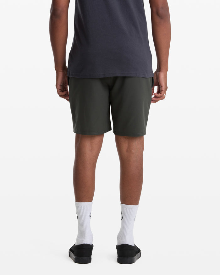 A person faces away, wearing a dark t-shirt, Volcom Men's Nomoly Elastic Waist Short 18 in dark green, white crew socks, and black slip-on shoes against a plain white background.