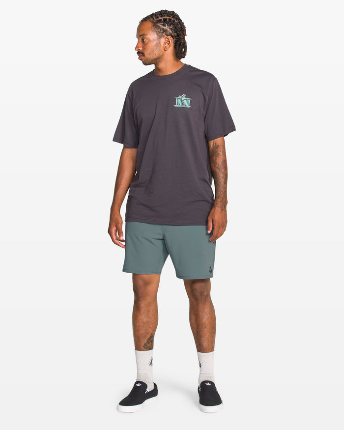 A man with braided hair stands facing left, wearing a black t-shirt with a small turquoise graphic, Volcom Men's Nomoly Elastic Waist Shorts 18" in teal made from recycled polyester, white socks, and black slip-on shoes against a white background.