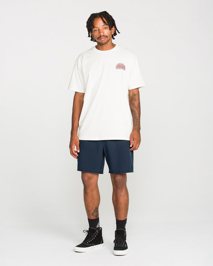 A man stands against a plain white background, wearing a white t-shirt with a small graphic on the chest and Volcom Men's Nomoly Elastic Waist Shorts in navy, which are made from a recycled blend. He completes his look with black socks and black high-top sneakers. He has tattoos on his arms and short, twisted hair.