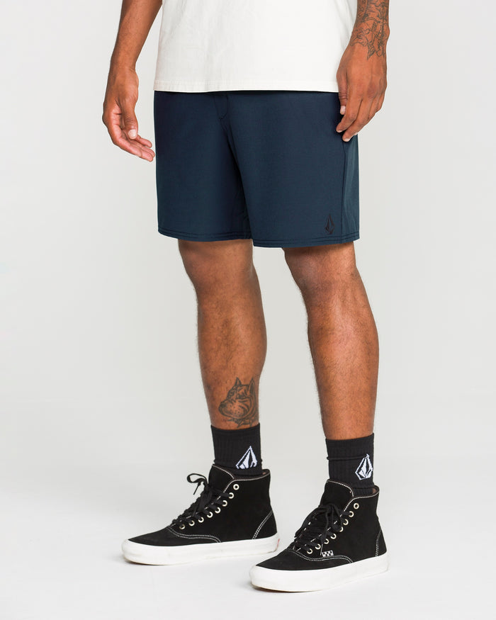 A person is wearing a white t-shirt, Volcom Men's Nomoly Elastic Waist Shorts in dark blue with 4-way stretch and made from a recycled blend, black high-top sneakers, and black socks with a white logo, standing against a plain white background.