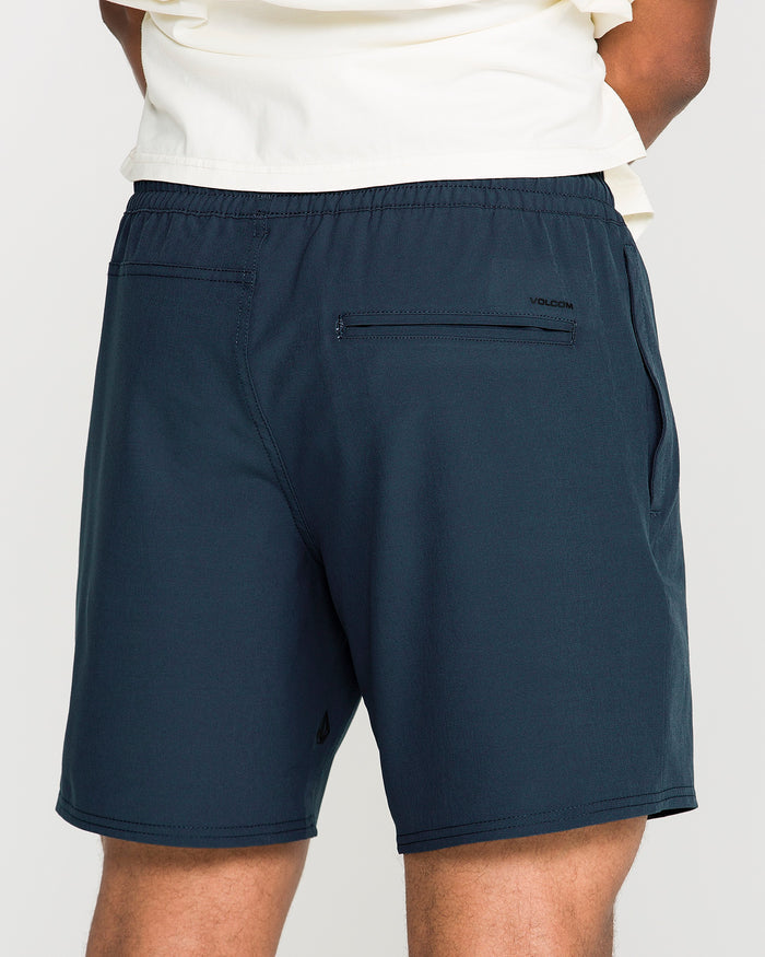 Back view of a person wearing a cream-colored shirt and Volcom Men's Nomoly Elastic Waist Shorts in navy blue, featuring side and back pockets, standing with hands behind their back against a plain background.