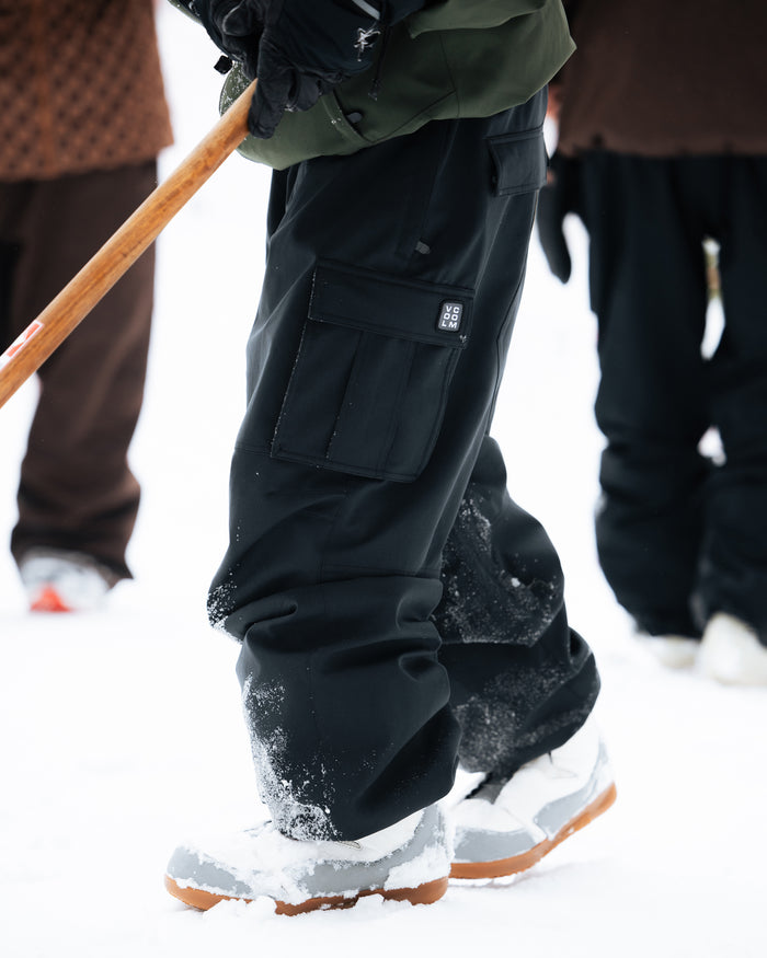 A person wearing Volcom Men’s Nwrk 20K Baggy Pants—crafted from a 20K waterproof, recycled shell—and gray boots stands in the snow holding a wooden stick, with others visible in the snowy background.