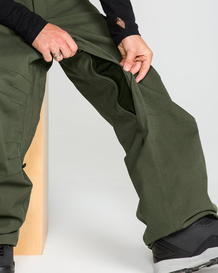 A person sits on a wooden bench, unzipping the side of olive green Volcom Men’s Nwrk 20K Baggy Pants to reveal the interior, with only their lower body and hands in view.