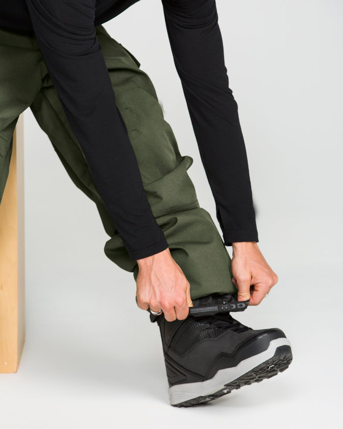 A person wearing Volcom Men’s Nwrk 20K Baggy Pants in green and a black long-sleeve shirt is tightening the strap on a black and gray winter boot. Only their lower body and hands are visible, drawing attention to the pants’ durable 20K waterproof fabric.