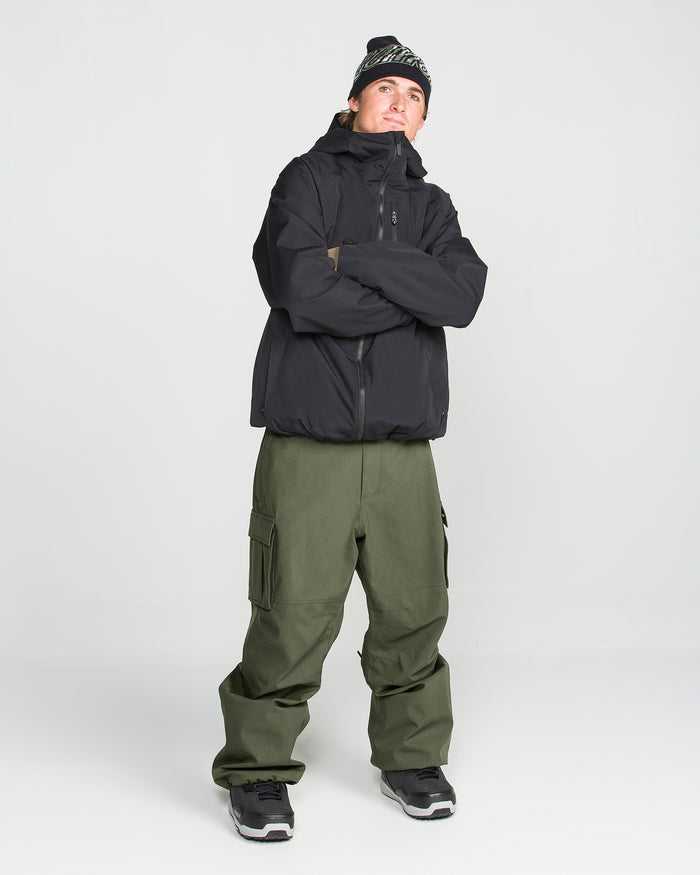 A person stands with arms crossed, looking slightly up, against a plain white background. They are wearing a black jacket, black sneakers, a black beanie, and Volcom Men’s Nwrk 20K Baggy Pants in olive green.