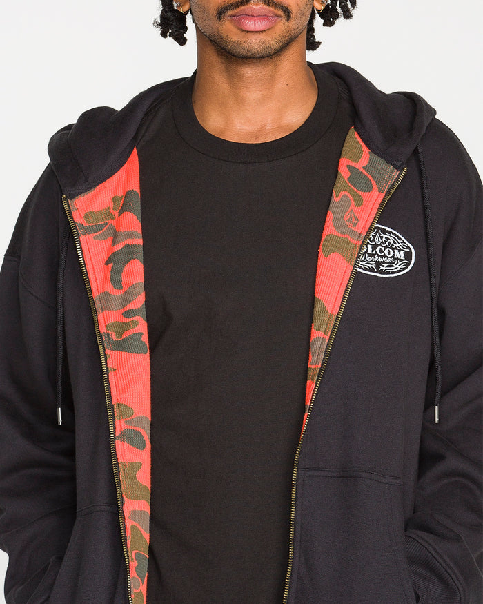 A person wearing Volcom’s Men’s On The Hunt Workwear Zip—a black hoodie with a red camo-patterned lining made from recycled polyester—along with a black t-shirt. Only the lower half of their face is visible.