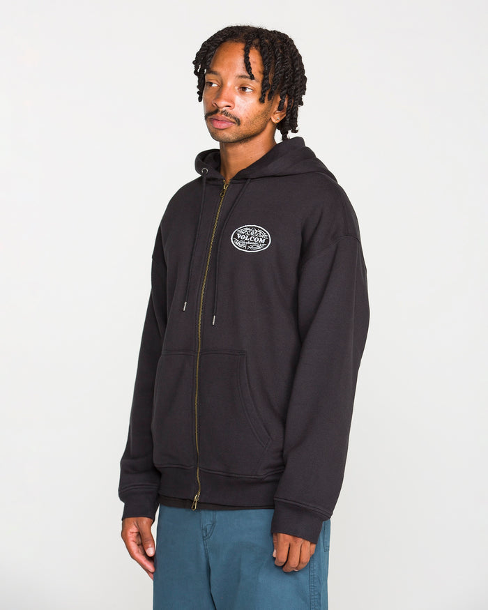 A man is wearing the Volcom Men’s On The Hunt Workwear Zip—a black fleece hoodie with a cotton waffle thermal lining and a circular chest logo—paired with blue pants, standing against a plain white background.