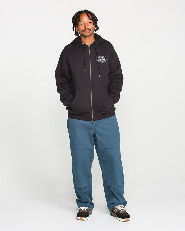 A man with short dreadlocks wears the Volcom Men’s On The Hunt Workwear Zip, a black fleece hoodie with a round logo and cotton waffle thermal lining. He pairs it with blue cargo-style pants made from recycled polyester and black sneakers, standing facing forward with his hands in his pockets against a plain white background.