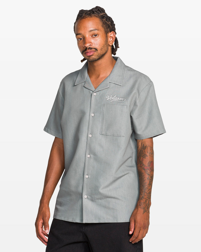 A man with tattoos on his right arm and braided hair wears the Volcom Men's Otis Stone Short Sleeve Tee, a boxy fit, textured light gray shirt with "Welcome" embroidered on the chest pocket. He stands against a plain white background.