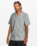 A man with tattoos on his right arm and braided hair wears the Volcom Men's Otis Stone Short Sleeve Tee, a boxy fit, textured light gray shirt with "Welcome" embroidered on the chest pocket. He stands against a plain white background.