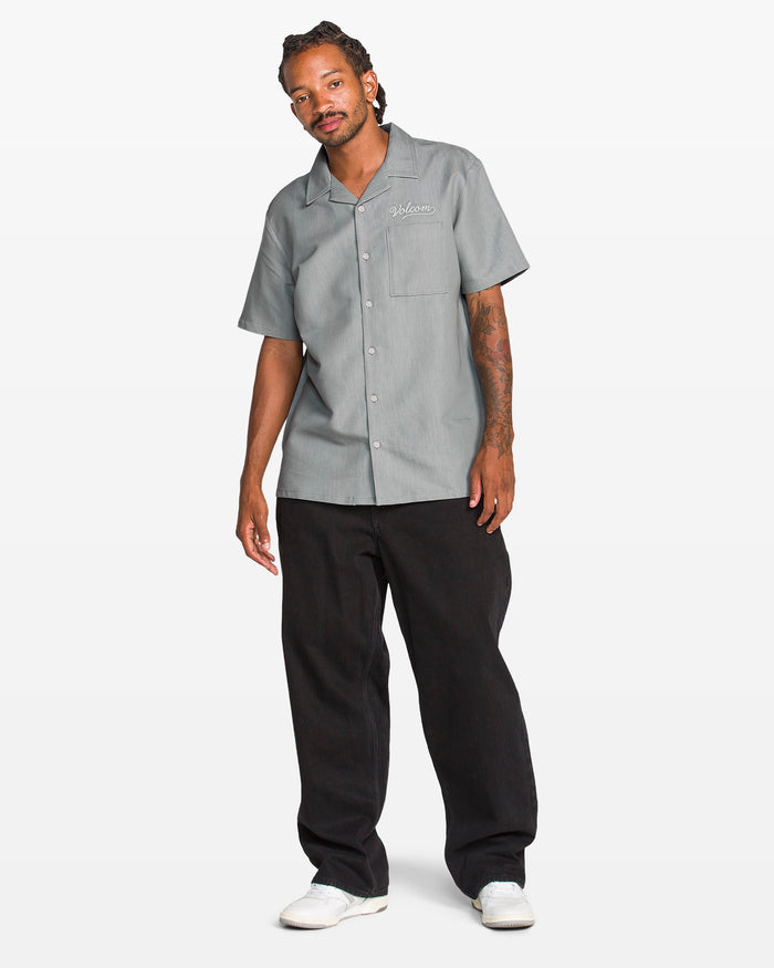 A man wears the Volcom Men's Otis Stone Short Sleeve Tee with black pants and white shoes, standing against a plain white background. He has tattoos on his right forearm and looks slightly to the left.