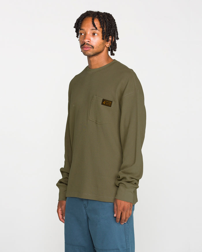 A man wearing a Volcom Men's Overtime Workwear Thermal Long Sleeve Shirt in olive green, featuring a chest pocket, and blue pants stands against a plain white background. Made from a cotton knit fabric, this thermal shirt provides comfort and durability for everyday wear.