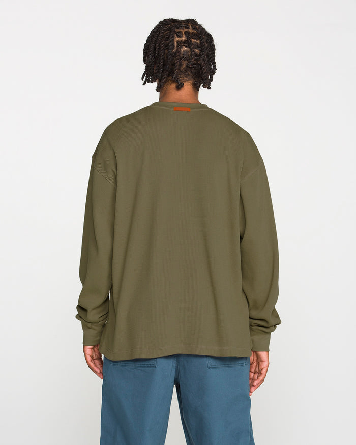A person with braided hair, wearing the Volcom Men's Overtime Workwear Thermal Long Sleeve Shirt in olive green and blue pants, stands facing away from the camera against a plain white background.