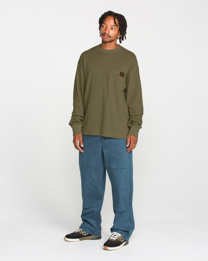 A person stands against a white background wearing a Volcom Men's Overtime Workwear Thermal Long Sleeve Shirt in olive green, blue pants, and black sneakers with white soles.