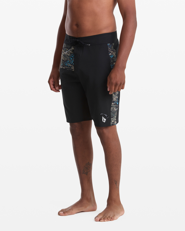 A man stands barefoot wearing Volcom Men's Parillo Mod 21 black board shorts with a drawstring waist and patterned side panels. His upper body is unclothed, showing tattoos on his left thigh and right ankle.