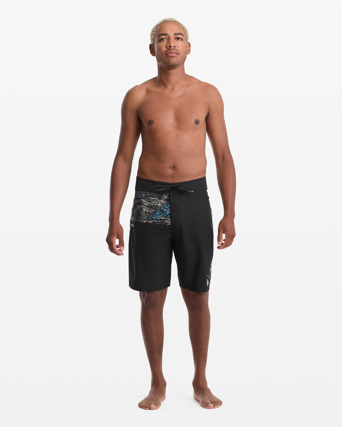A man with short, bleached hair stands barefoot against a white background, wearing Volcom Men's Parillo Mod 21 black board shorts with a patterned side. He is shirtless, facing forward with a neutral expression.
