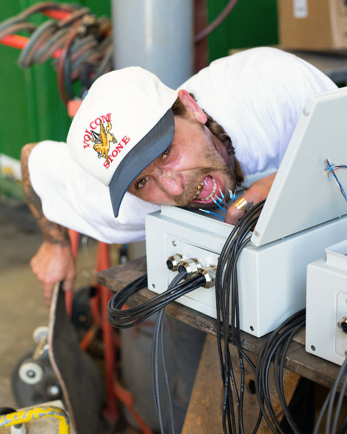 A man wearing a Volcom Men's Petrolstone Adj Hat and t-shirt leans over an electrical box with wires in his mouth, holding a skateboard amidst cables and industrial equipment.